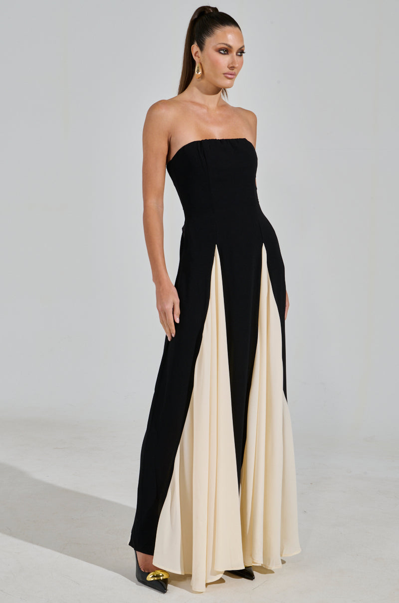 A woman confidently stands in the ON MY FYP CHIFFON MAXI DRESS, featuring a strapless silhouette with cream panels. Her long hair is tied back, and she accessorizes with drop earrings and black shoes with gold tips. The minimalist background highlights her elegant attire.