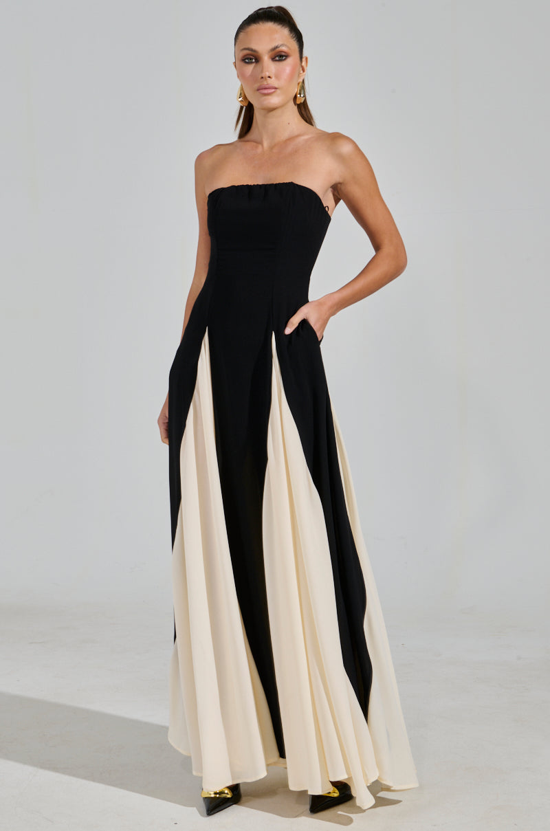 A model elegantly showcases the ON MY FYP CHIFFON MAXI DRESS, featuring a strapless silhouette with a black top and a flowing black and cream skirt. Posing against a light gray backdrop, she accessorizes with gold earrings and black shoes, exuding timeless style.