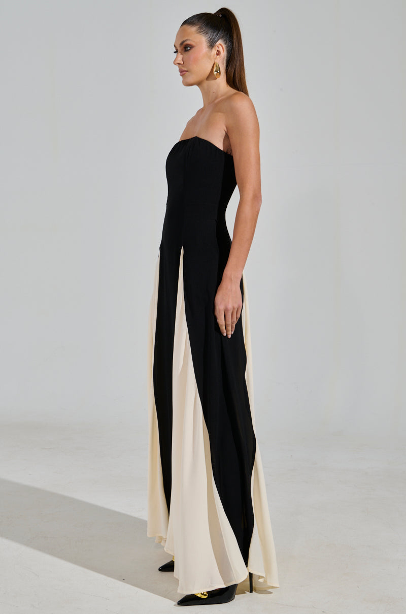 A woman poses in profile, showcasing the ON MY FYP CHIFFON MAXI DRESS—a black and white strapless design with flowing chiffon panels. She pairs it with large gold earrings, a high ponytail, and elegant black high heels.