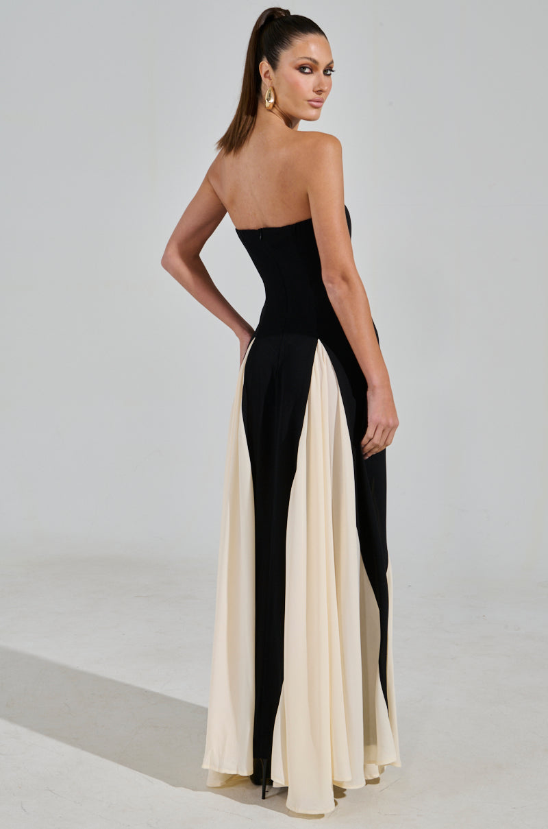 A woman models the ON MY FYP CHIFFON MAXI DRESS in a black strapless silhouette with white flowing panels, standing sideways and looking over her shoulder, her hair styled in a high ponytail.