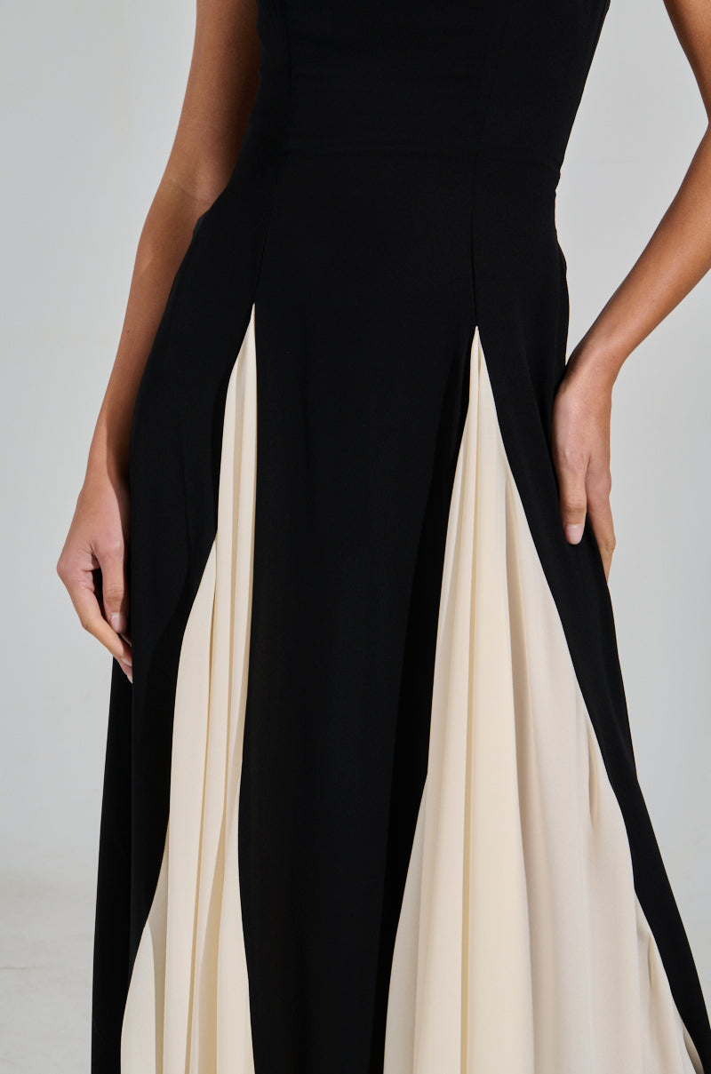 Wearing the "ON MY FYP CHIFFON MAXI DRESS," a person poses against a neutral backdrop. The black chiffon maxi dress flaunts cream-colored pleats and an A-line design with contrasting panels. Only the torso and upper legs are visible, with one hand resting on the hip.