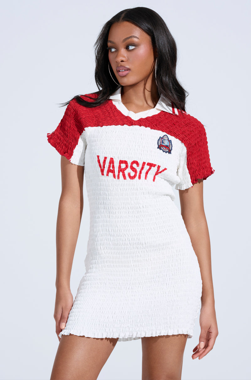 Wearing the TEAM PLAYER SMOCKED JERSEY DRESS, featuring a bold red and white color block and "VARSITY" with a sports patch, a person with long dark hair looks to the side against a plain white backdrop.
