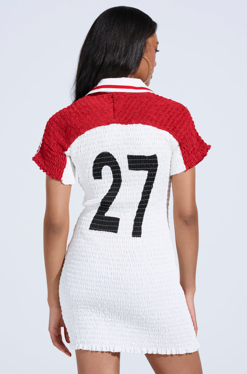 Wearing a red and white TEAM PLAYER SMOCKED JERSEY DRESS with a number 27 patch on the back, a person with long hair stands facing away against a plain background.