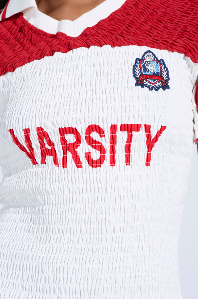 Close-up of the TEAM PLAYER SMOCKED JERSEY DRESS, featuring red and white color blocks with "VARSITY" in bold red letters across the chest. An embroidered crown patch with decorative details on the upper left adds an athletic touch to this classic design.