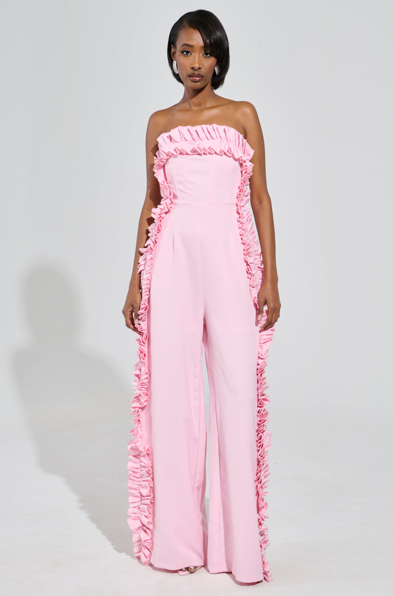 A woman stands confidently in the MONICA RUFFLED JUMPSUIT IN PINK, featuring a strapless neckline and ruffled detailing down the sides, complemented by a wide leg silhouette. The plain white background emphasizes the elegance and modern style of her outfit.