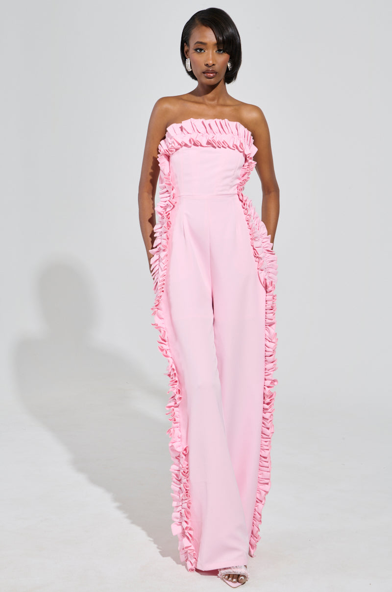 A person confidently poses against a plain white background, wearing the MONICA RUFFLED JUMPSUIT IN PINK, which features a strapless neckline and wide leg silhouette, with one hand in their pocket.