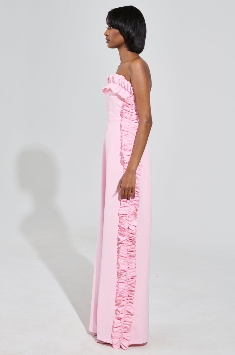 A woman in a long, pink MONICA RUFFLED JUMPSUIT with a strapless neckline stands against a plain background. She is facing sideways, with her arms by her sides, showcasing the elegant details of the jumpsuit.