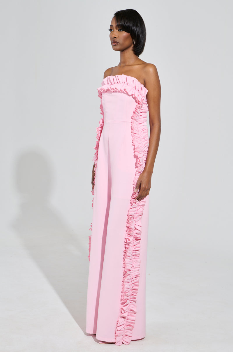 A woman is wearing the MONICA RUFFLED JUMPSUIT IN PINK, featuring a wide-leg design, strapless neckline, and ruffled trim along the top and sides. She stands against a plain white background, looking to the side.