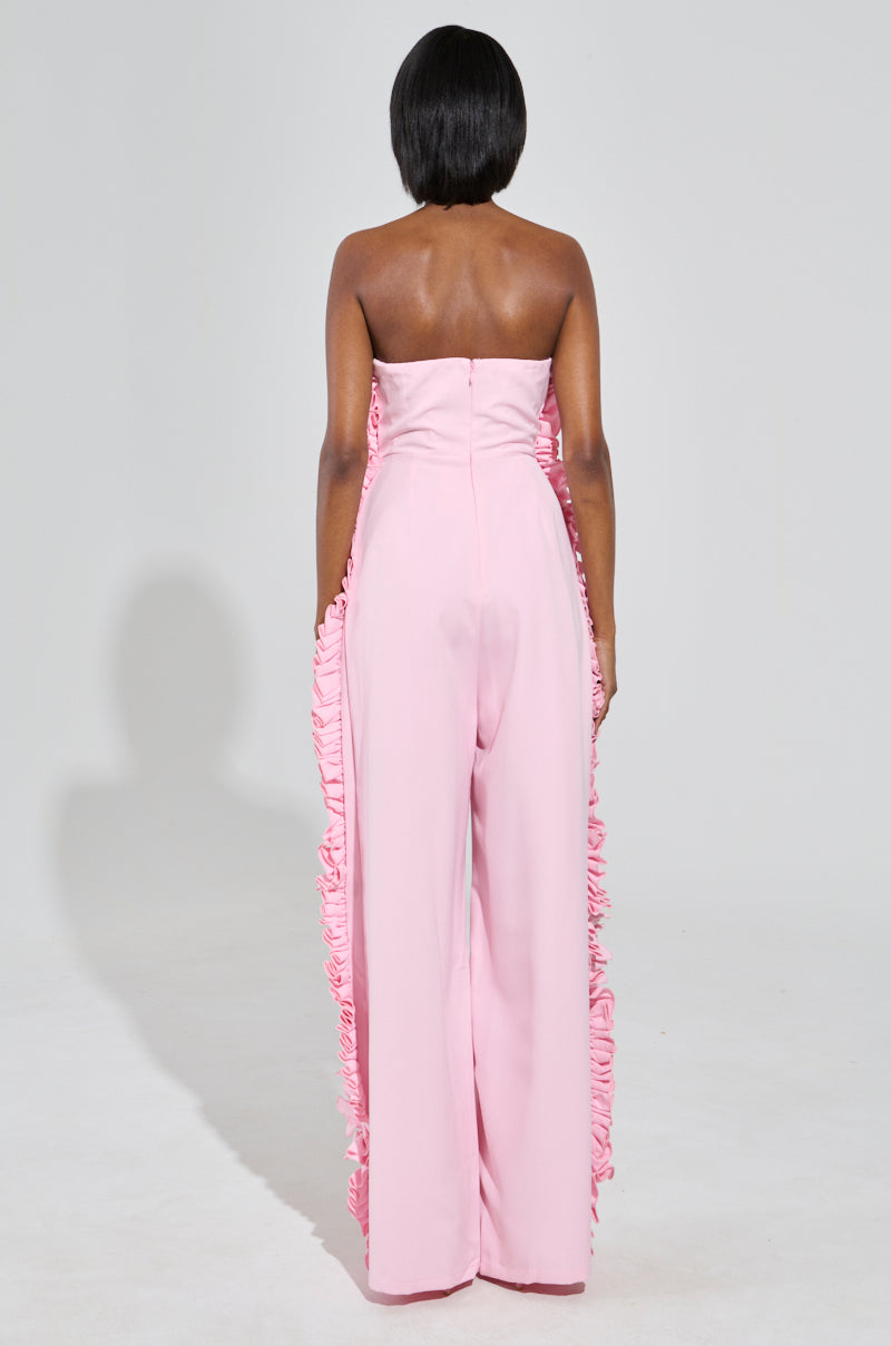 A person stands facing away, dressed in the MONICA RUFFLED JUMPSUIT IN PINK, which features a strapless neckline and a wide-leg silhouette against a plain white background.