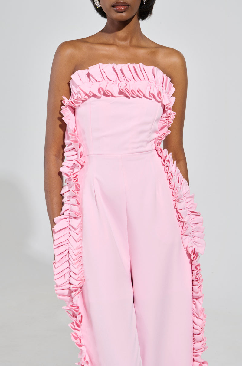 A woman is wearing the MONICA RUFFLED JUMPSUIT IN PINK with a wide-leg silhouette and strapless neckline. The plain background highlights the outfit's elegant design.