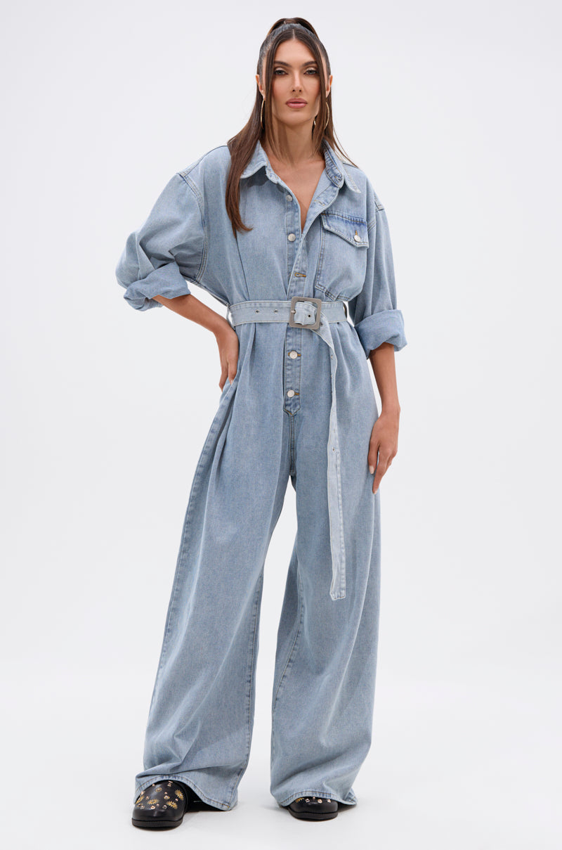A woman poses confidently against a white backdrop, wearing the NOT TODAY EXTREME OVERSIZED DENIM JUMPSUIT with a matching belt, black shoes, and straight hair styled down.