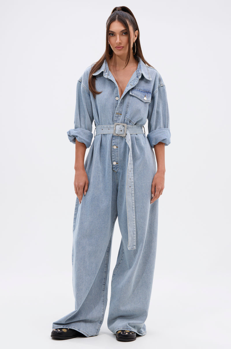 A woman stands facing forward in the NOT TODAY EXTREME OVERSIZED DENIM JUMPSUIT, featuring a light wash, adjustable waist belt, wide legs, and rolled sleeves. She wears hoop earrings and black shoes against a plain white background.