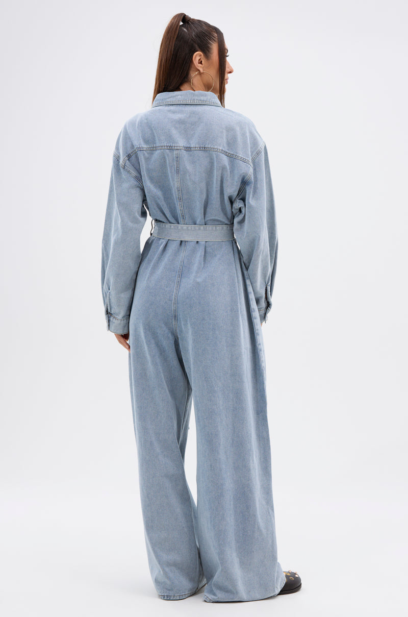 A woman with long brown hair in a ponytail faces away, wearing the NOT TODAY EXTREME OVERSIZED DENIM JUMPSUIT with an adjustable waist belt, long sleeves, and wide legs, set against a plain white background.