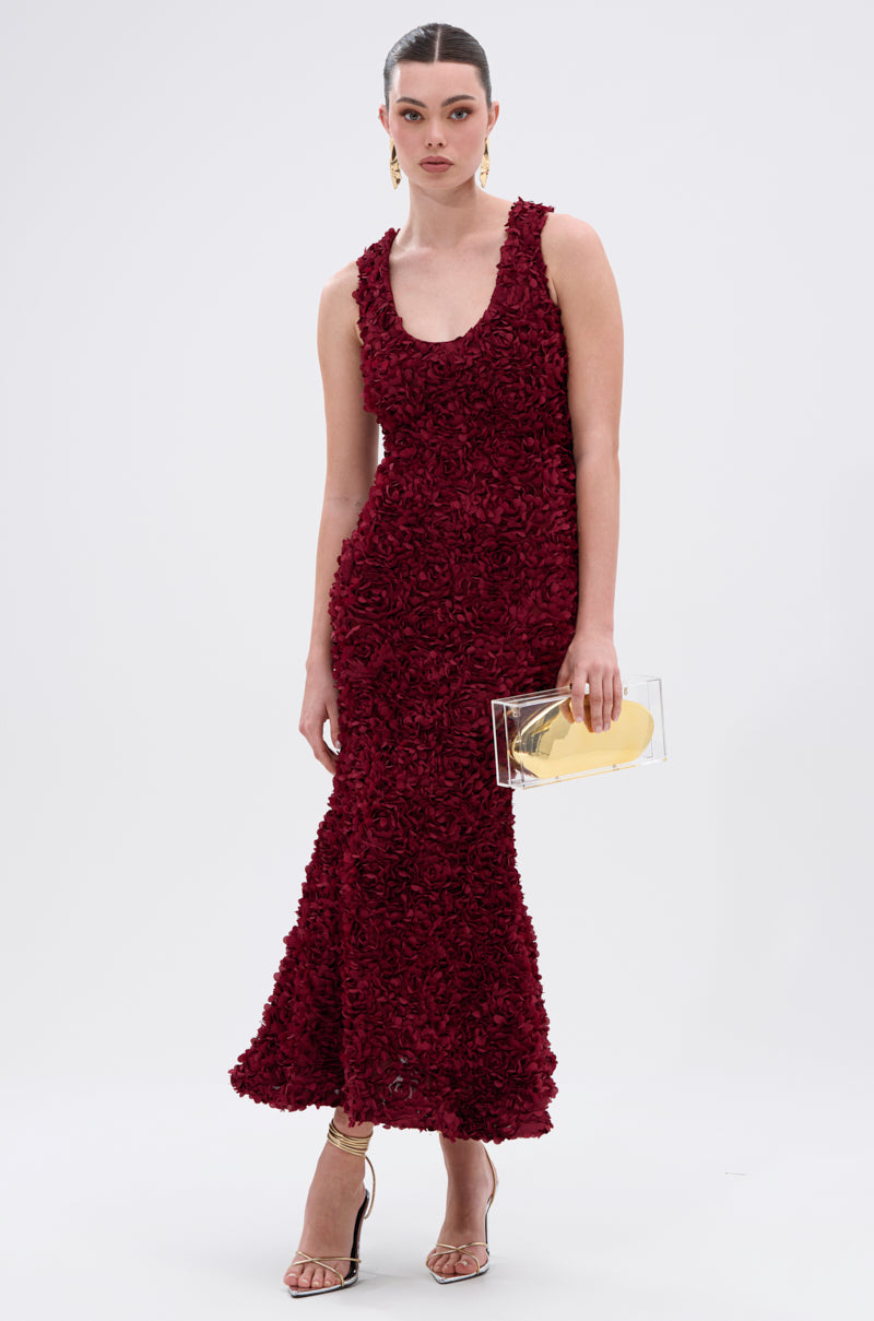 A woman wears the GARDEN PARTY FLORAL MIDI DRESS, styled with gold earrings, strappy heels, and a transparent clutch with a gold accent. Her hair is pulled back, and she stands against a plain white background.