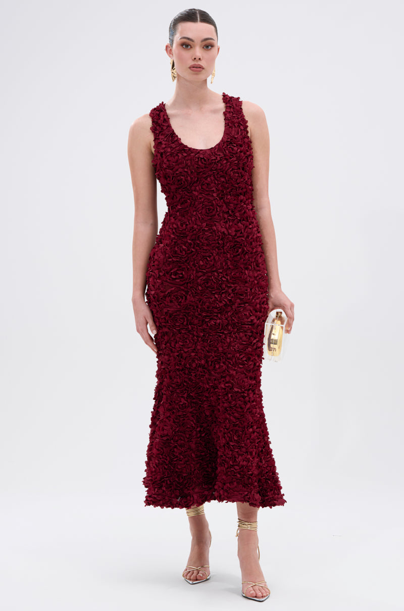 A woman poses in the GARDEN PARTY FLORAL MIDI DRESS—a sleeveless, fitted wine-red dress with a floral texture and mermaid silhouette. She accessorizes with a gold clutch, strappy gold heels, and wears her hair in a sleek bun.
