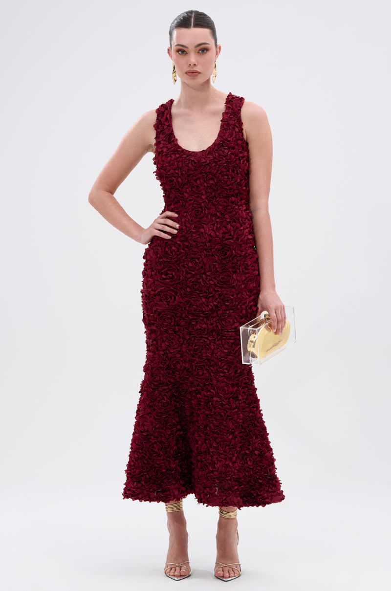 A woman stands confidently in the GARDEN PARTY FLORAL MIDI DRESS, a wine-red mermaid silhouette. She holds a transparent clutch with gold accent, accessorized with gold earrings and strappy heels against a plain white background.