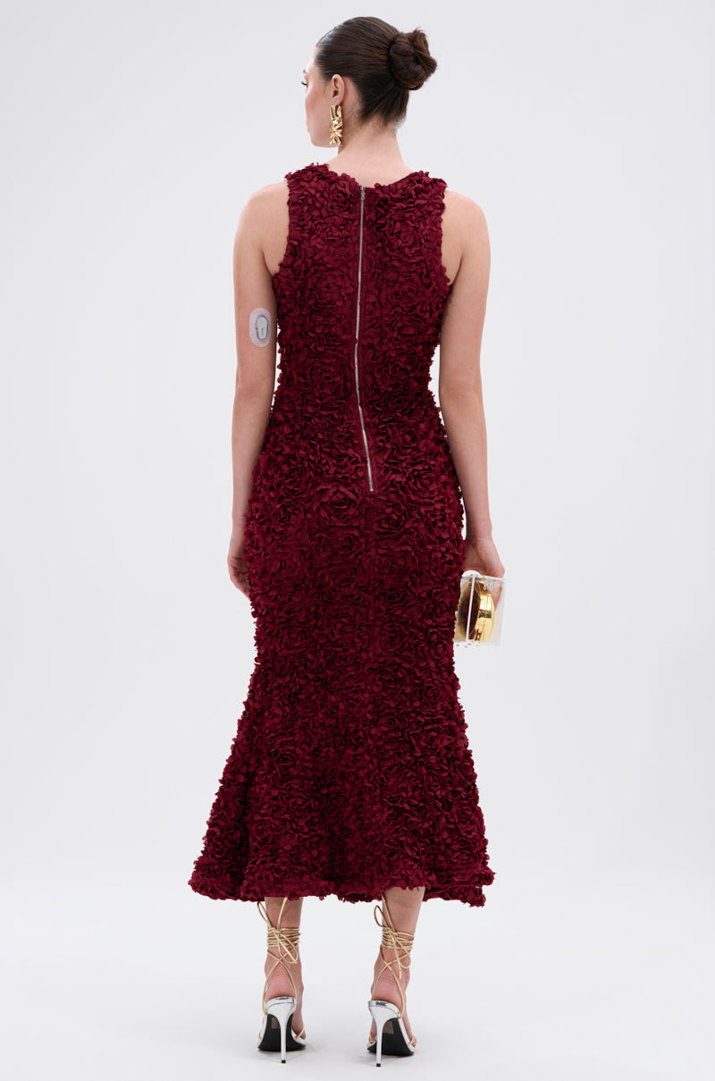 A woman in the GARDEN PARTY FLORAL MIDI DRESS, featuring a textured, sleeveless wine-red design, faces away. She holds a gold clutch, wears gold strappy heels and earrings, hair in a bun, with a small patch on her upper arm.
