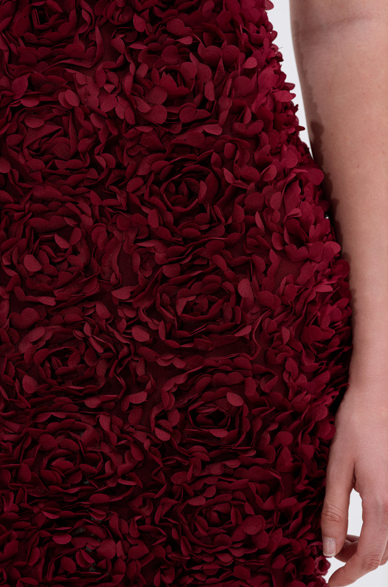 A close-up of a person wearing the GARDEN PARTY FLORAL MIDI DRESS in wine-red, adorned with textured 3D floral patterns. The person’s arm and hand appear on the right side of the image.