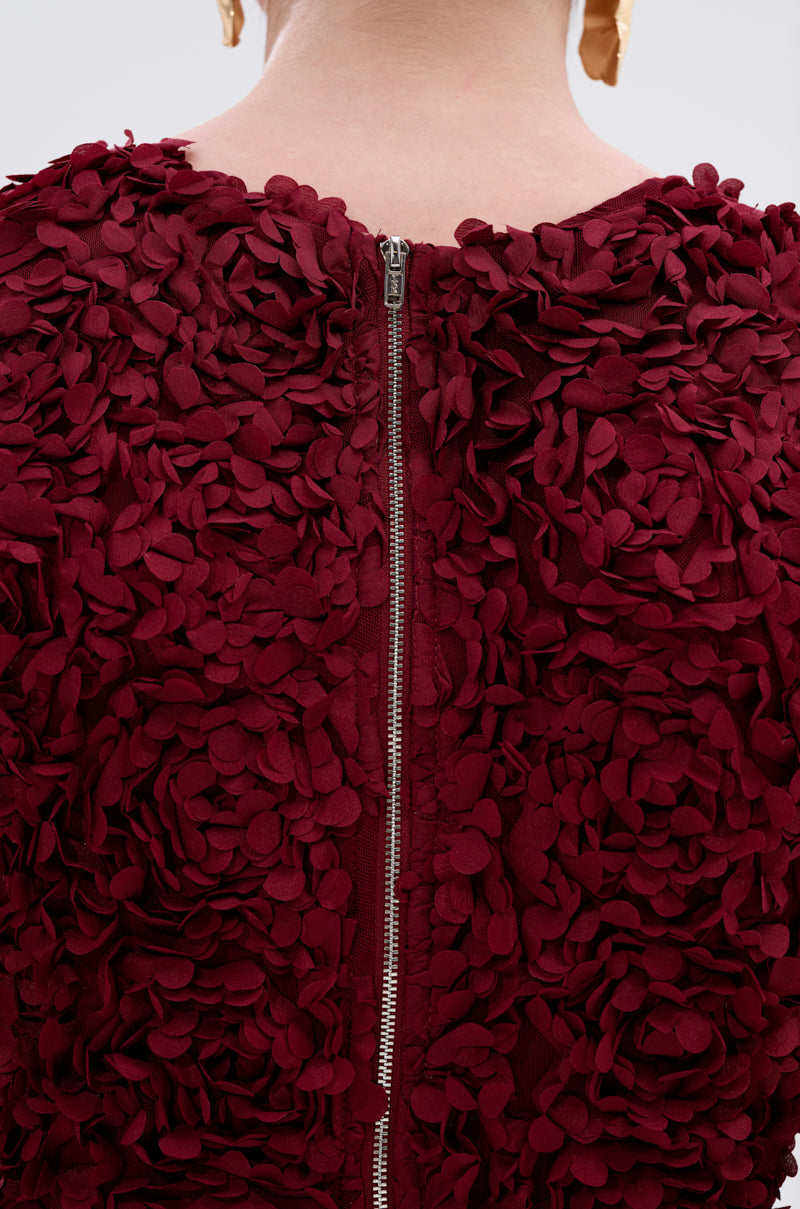 Close-up of a person in the GARDEN PARTY FLORAL MIDI DRESS, featuring wine-red petal-like details and a silver back zipper; their neck and gold earrings are visible, highlighting the dress’s elegant, floral-inspired design.