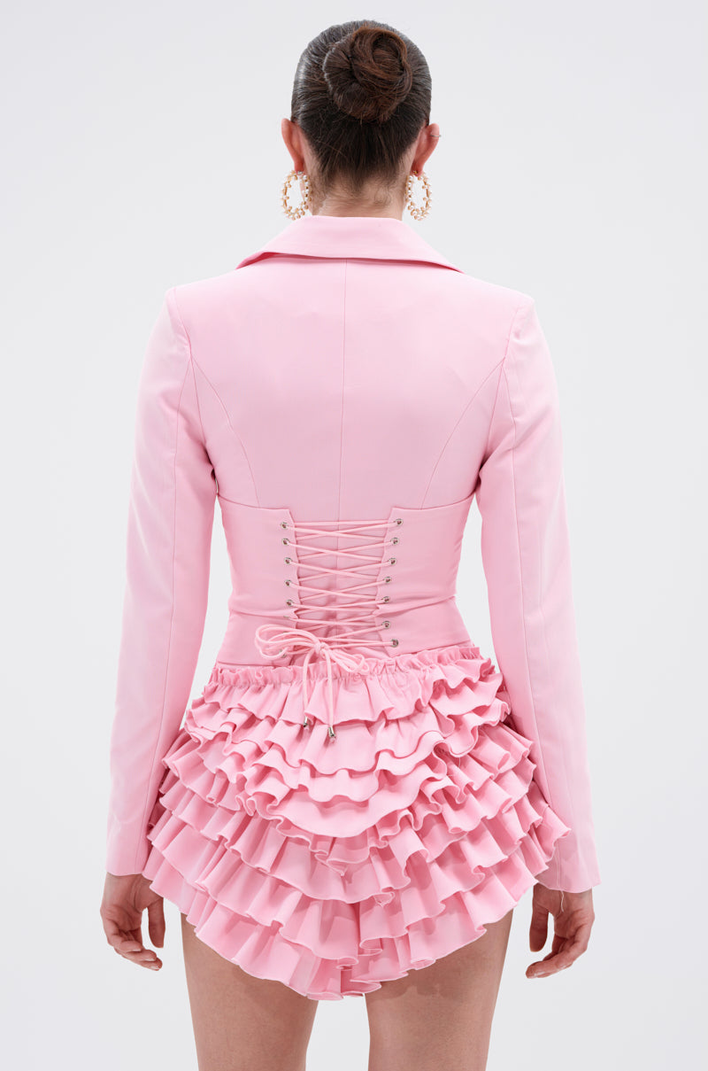 A woman with a neat bun is seen from behind wearing the STRICTLY BUSINESS BLAZER ROMPER IN PINK, featuring a lace-up corset detail and tiered ruffle skirt, against a plain light background.
