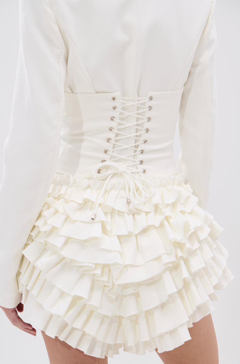 Wearing the STRICTLY BUSINESS BLAZER ROMPER IN WHITE, a person is seen from the back, showcasing its fitted lace-up corset-style back, notched lapel collar, and double-layered ruffle hem.