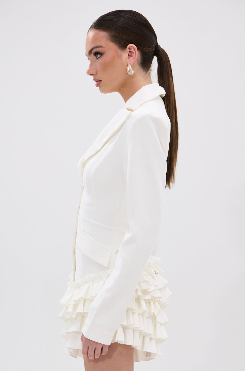 A woman with long, straight brown hair in a low ponytail models the STRICTLY BUSINESS BLAZER ROMPER IN WHITE, featuring a notched lapel collar and double-layered ruffle skirt hem, posing in profile against a white background.