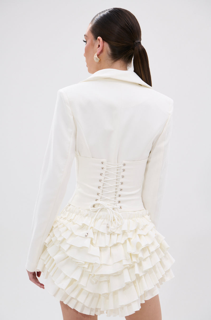 A woman with long brown hair in a ponytail wears the STRICTLY BUSINESS BLAZER ROMPER IN WHITE, featuring a notched lapel collar, double-layered ruffle hem, and lace-up back detail as she stands facing away from the camera.