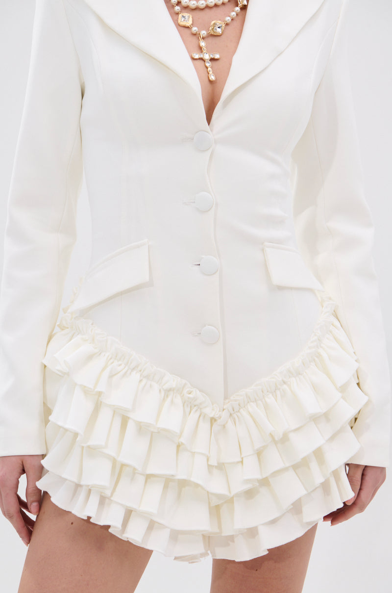 Wearing the STRICTLY BUSINESS BLAZER ROMPER IN WHITE, featuring a fitted silhouette, notched lapel collar, large buttons, flap pockets, and a double-layered ruffle hem, accessorized with a gold cross necklace.