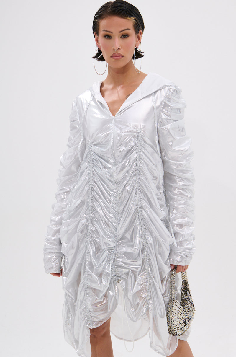 A woman stands against a plain white background wearing the WAYS TO GO METALLIC ADJUSTABLE DRESS IN WHITE, paired with a small snakeskin-patterned handbag, short slicked-back hair, and large hoop earrings.