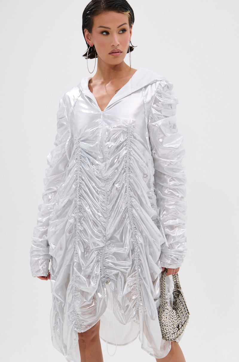 A woman models the WAYS TO GO METALLIC ADJUSTABLE DRESS IN WHITE—a shiny, ruched mini dress with long sleeves and a hood—paired with a silver textured handbag, hoop earrings, and sleek hair against a white background.