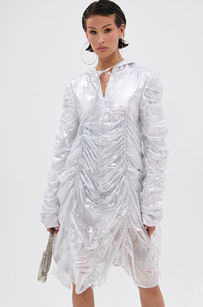 A person stands against a white background wearing the WAYS TO GO METALLIC ADJUSTABLE DRESS IN WHITE—a shiny, metallic ruched mini dress with long sleeves and a front slit—styled with large hoop earrings, short slicked-back hair, and a small beaded handbag.
