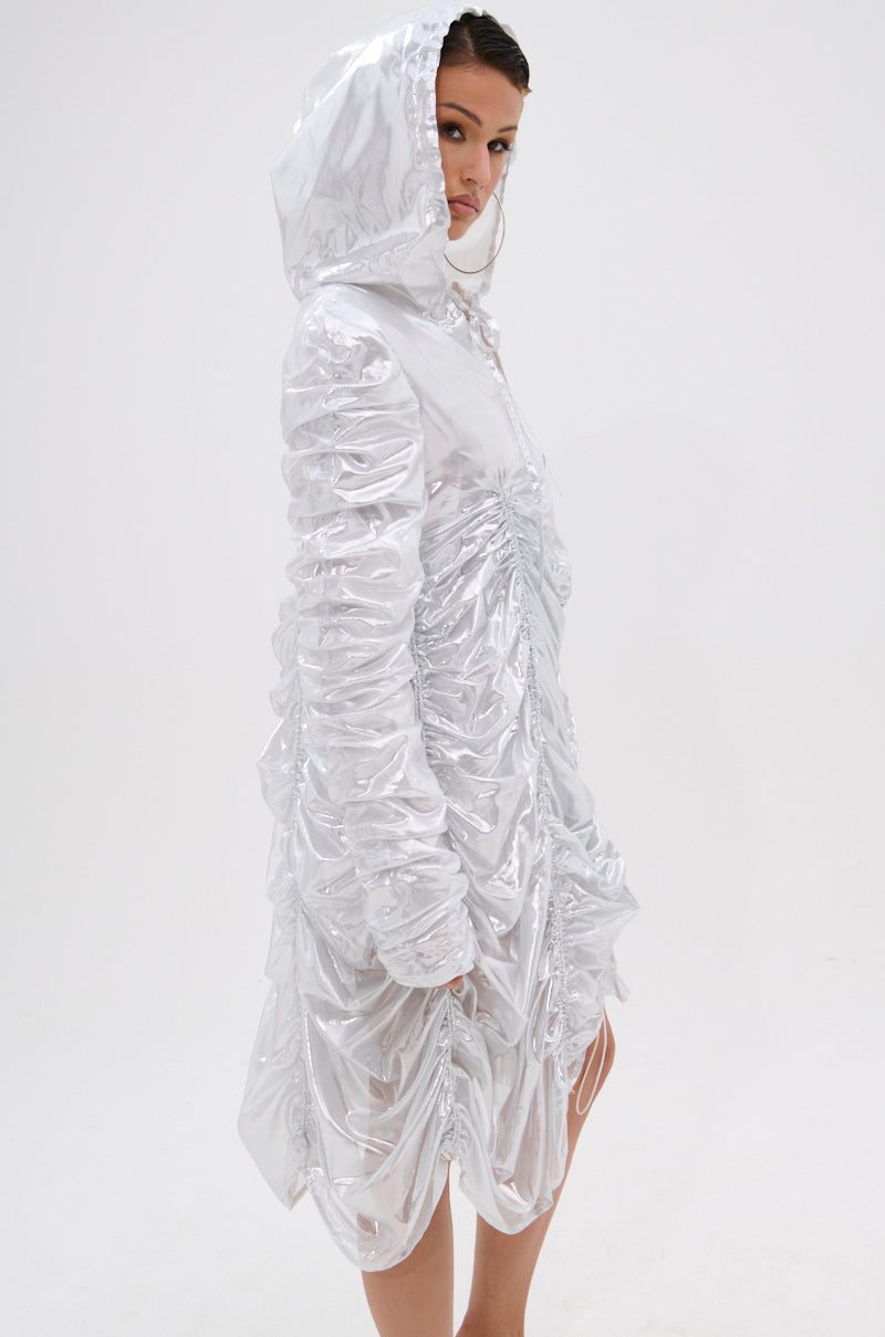 A woman models the WAYS TO GO METALLIC ADJUSTABLE DRESS IN WHITE—a hooded mini dress with ruching—standing against a plain white background, glancing over her shoulder with a neutral expression.