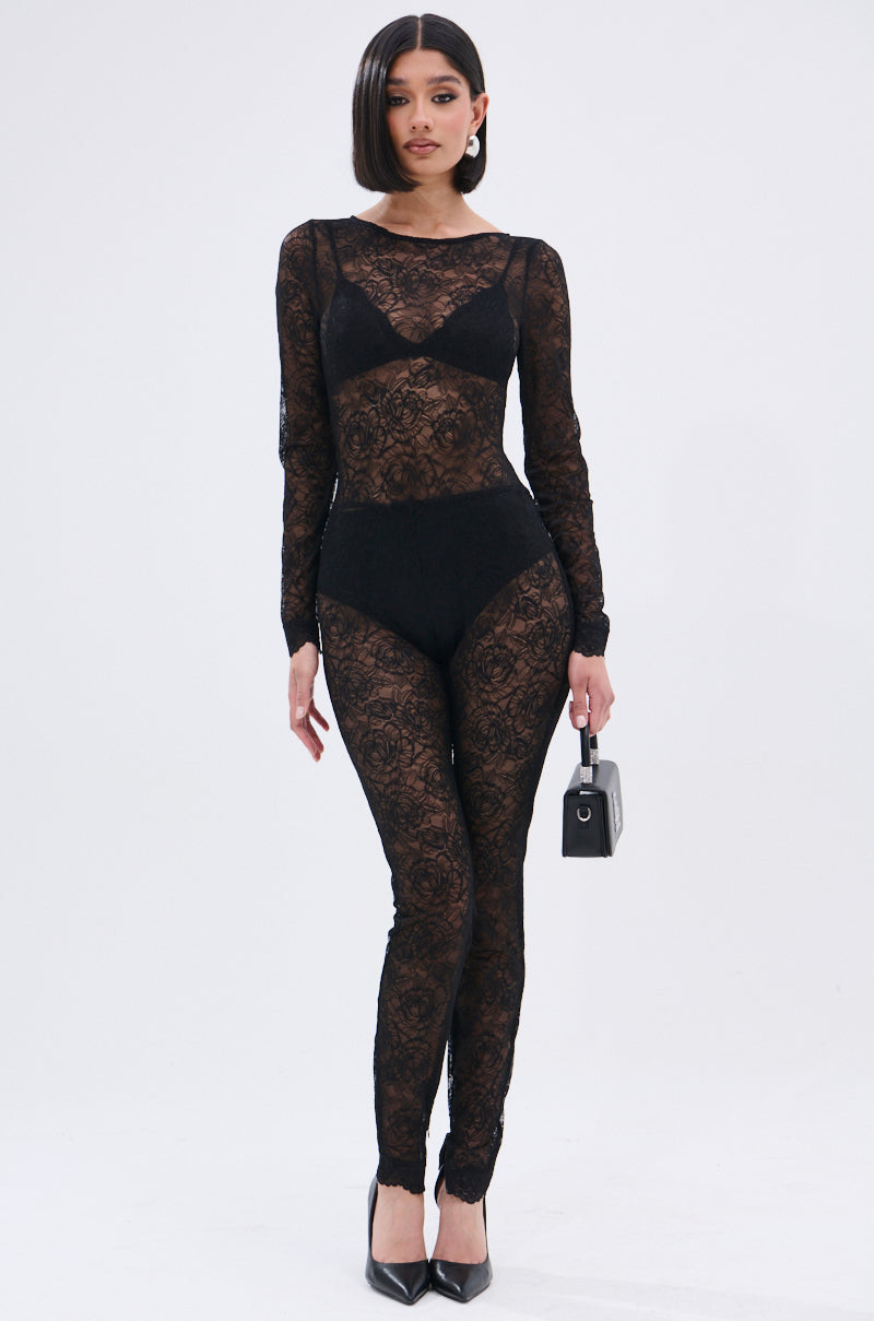 A woman poses against a plain backdrop in the ONLY IF THERE'S BOTTLE SERVICE LACE JUMPSUIT—curve-hugging, sheer black lace with floral details over black undergarments—styled with pointed black heels, a small black handbag, and chin-length straight dark hair.