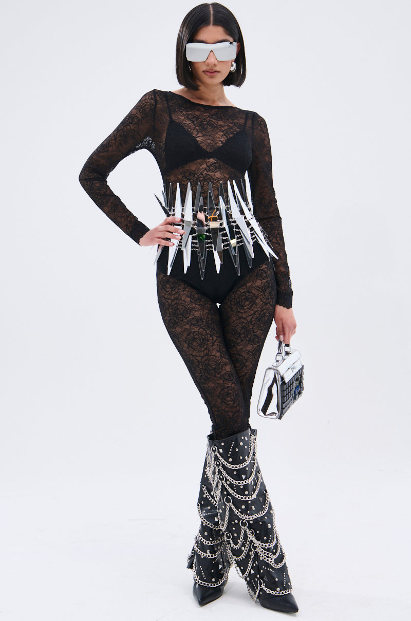 A woman models the ONLY IF THERE'S BOTTLE SERVICE LACE JUMPSUIT with black undergarments, mirrored visor sunglasses, a spiked belt, a small purse, and tall black boots adorned with chains and studs.