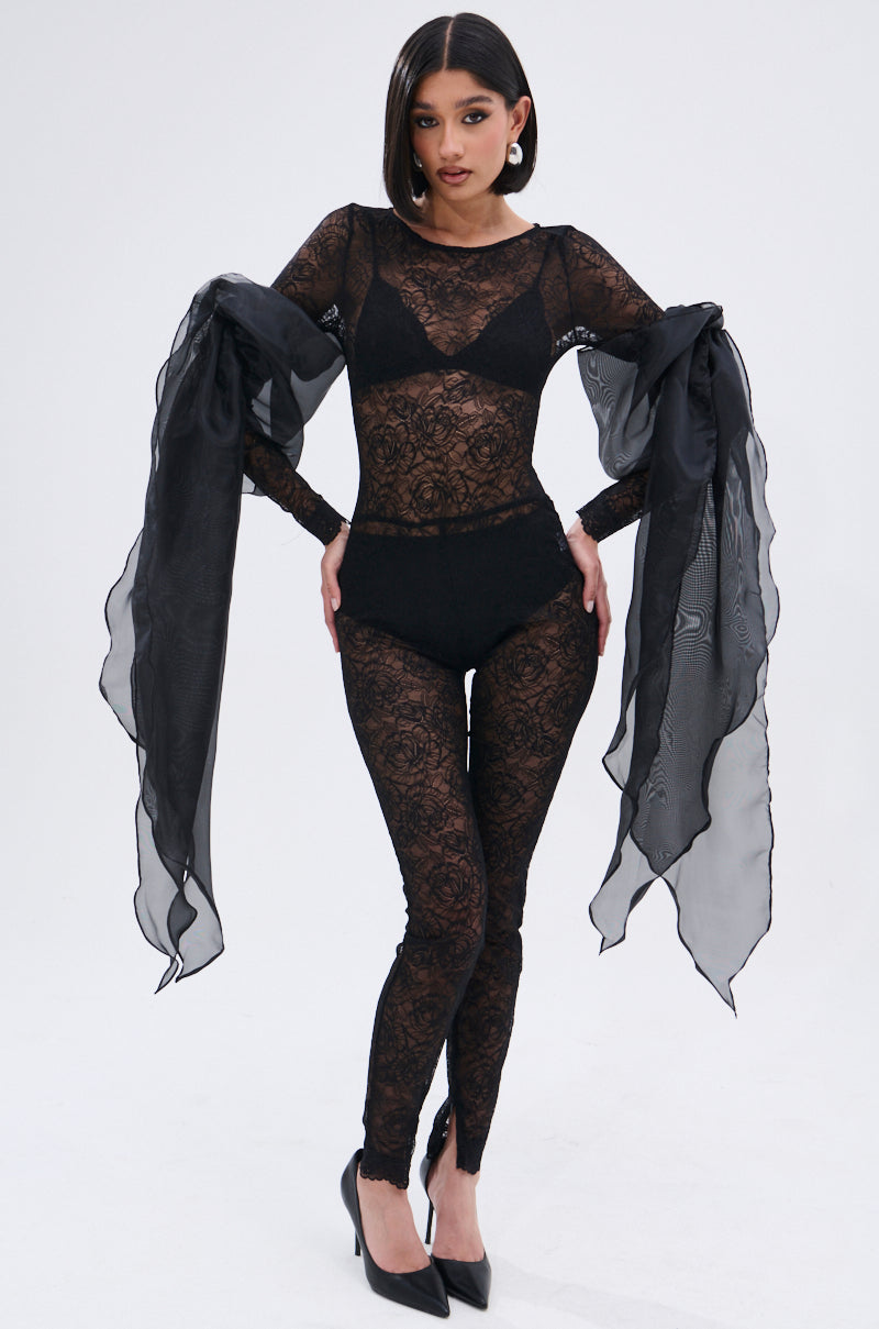 A woman poses confidently in the ONLY IF THERE'S BOTTLE SERVICE LACE JUMPSUIT, featuring sheer, curve-hugging lace and long chiffon bows on her sleeves. She stands in black heels against a plain, light background.