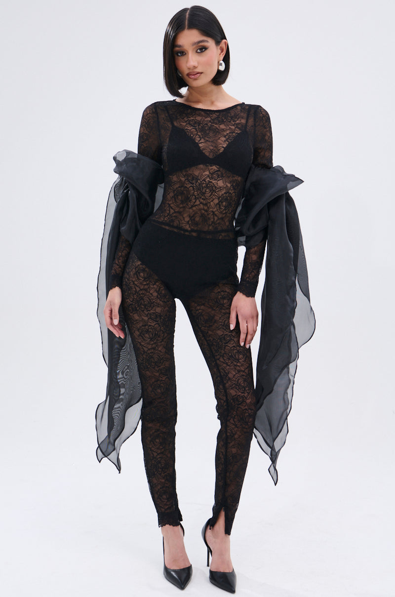A woman poses confidently in the ONLY IF THERE'S BOTTLE SERVICE LACE JUMPSUIT, featuring curve-hugging floral lace, long sleeves with dramatic tulle bows, and black high heels. She sports a sleek bob against a plain light background.