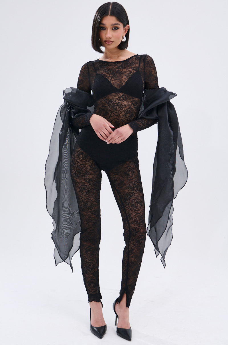 A woman models the ONLY IF THERE'S BOTTLE SERVICE LACE JUMPSUIT, a sheer black floral lace one-piece with a curve-hugging fit, layered over black lingerie and paired with high heels and a sheer shawl, against a plain white background.