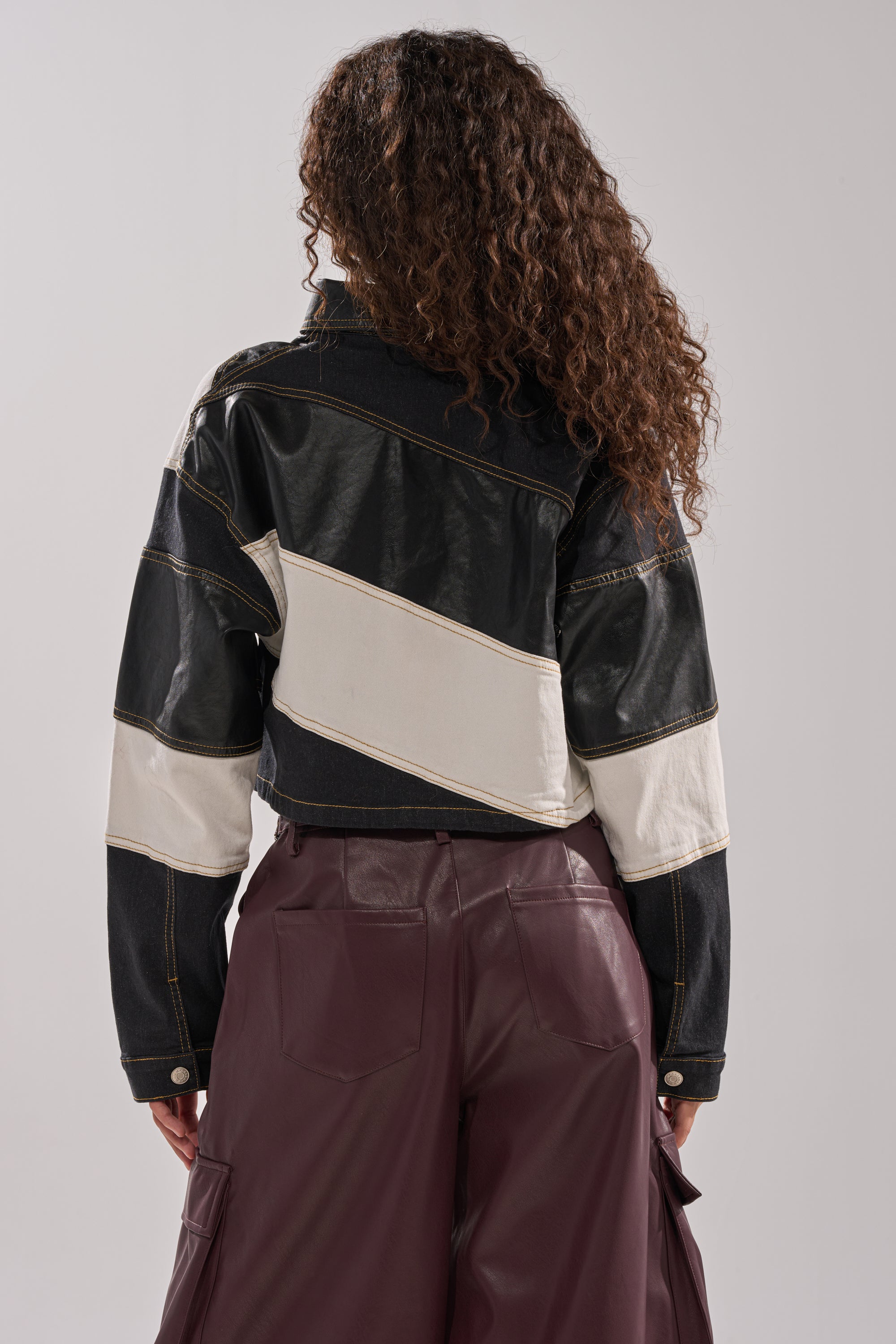 A person with curly hair stands facing away, wearing the GUESS WHAT STRIPE COLLARED BLOUSE. The top features black and white stripes and varsity-inspired details, paired with high-waisted maroon pants against a plain, light background.