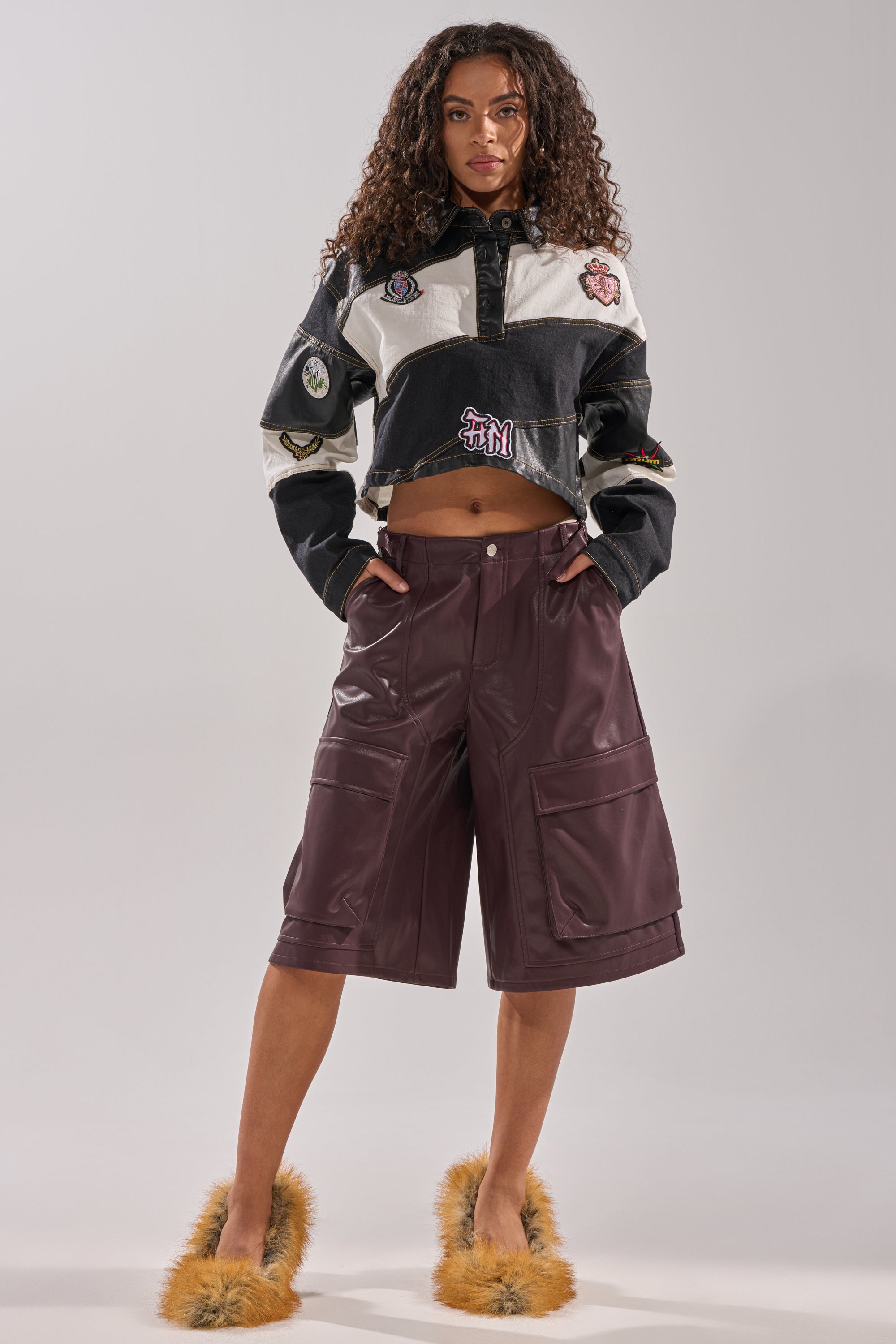 A woman stands confidently in the GUESS WHAT STRIPE COLLARED BLOUSE, paired with burgundy leather knee-length shorts with pockets and fuzzy slippers. She faces forward, hands in her pockets, against a plain background.