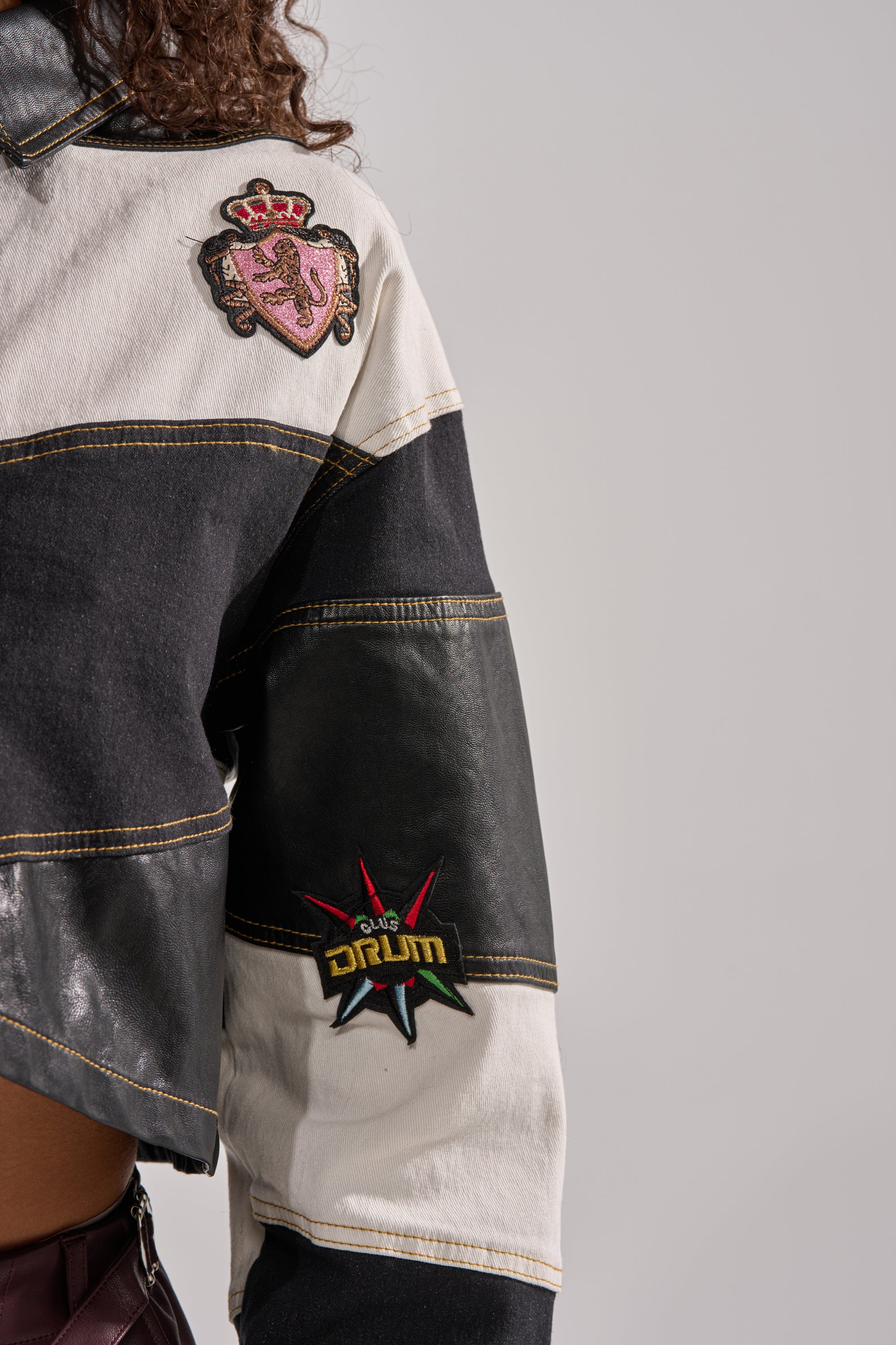 A person wears the GUESS WHAT STRIPE COLLARED BLOUSE, a black-and-white striped top decorated with varsity-style embroidered patches—a crest on the shoulder and a starburst with "DRUM" on the sleeve.