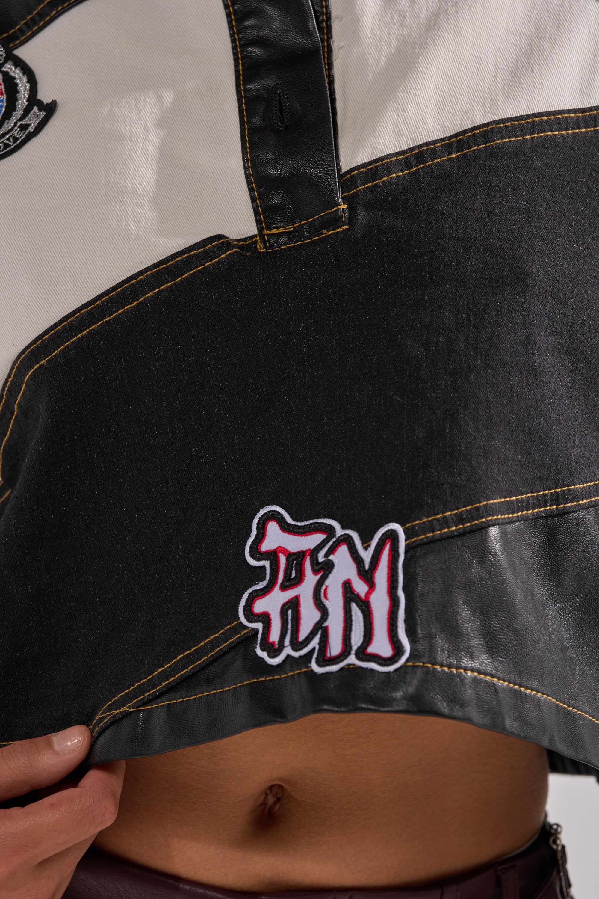 A close-up of someone lifting the hem of the GUESS WHAT STRIPE COLLARED BLOUSE, featuring black and white color-blocking, yellow stitching, varsity patches—including a red and white "NM." Their bare midriff and hand are partially visible.