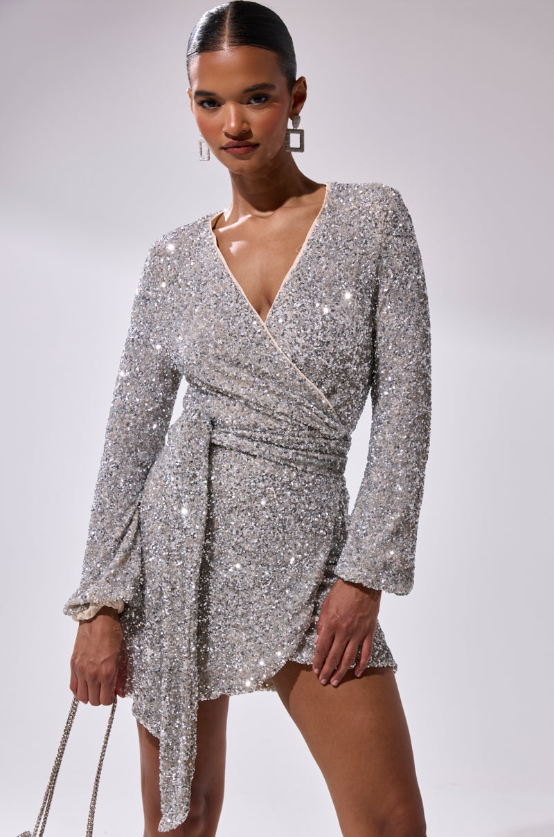 A woman models the LORETTA SEQUIN WRAP MINI DRESS with long sleeves and a deep V-neckline, pairing it with geometric earrings and a beige handbag against a plain light background.