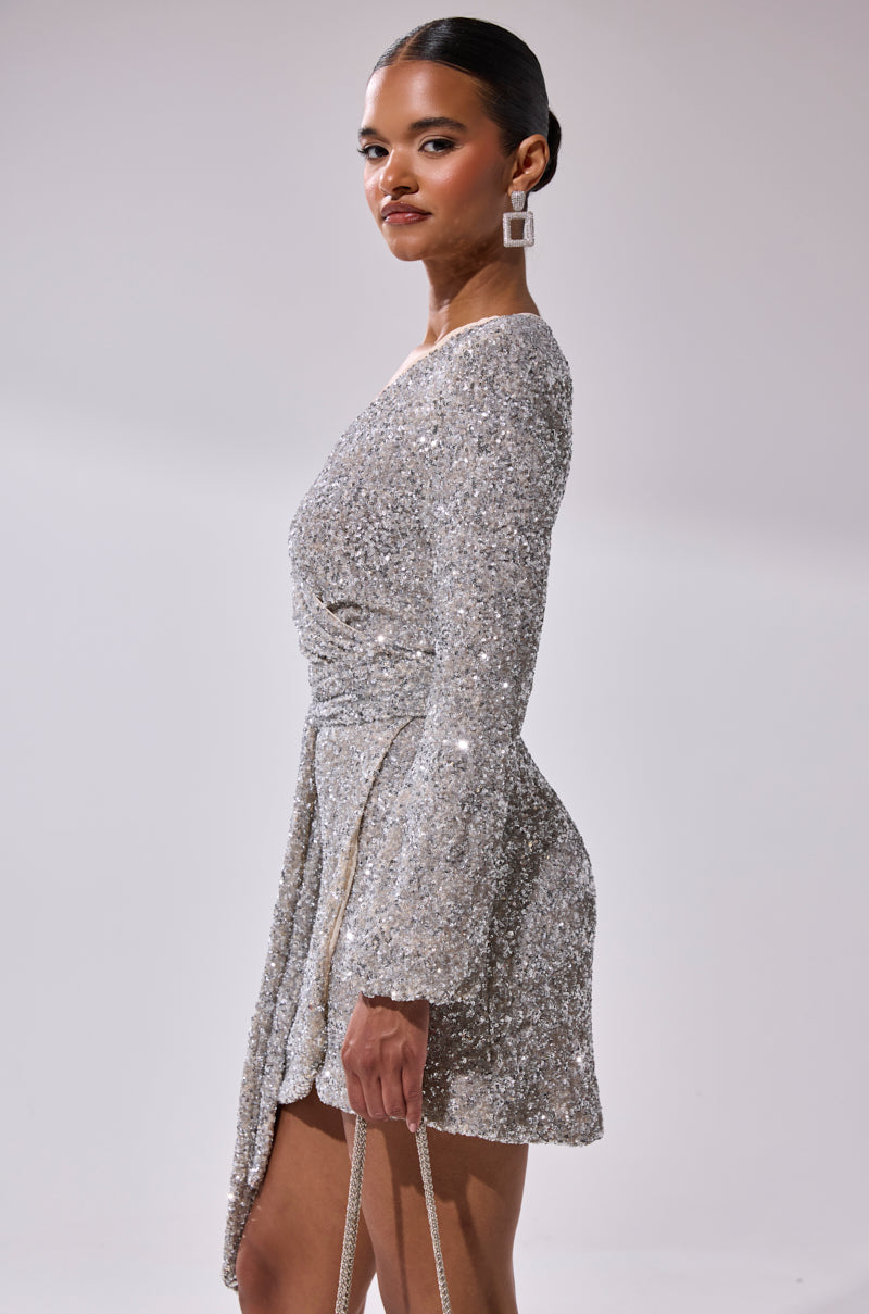 A woman poses confidently in the LORETTA SEQUIN WRAP MINI DRESS, a shimmering silver long-sleeve party dress. She completes her look with geometric earrings, a small bag, and pulled-back hair against a plain light background.