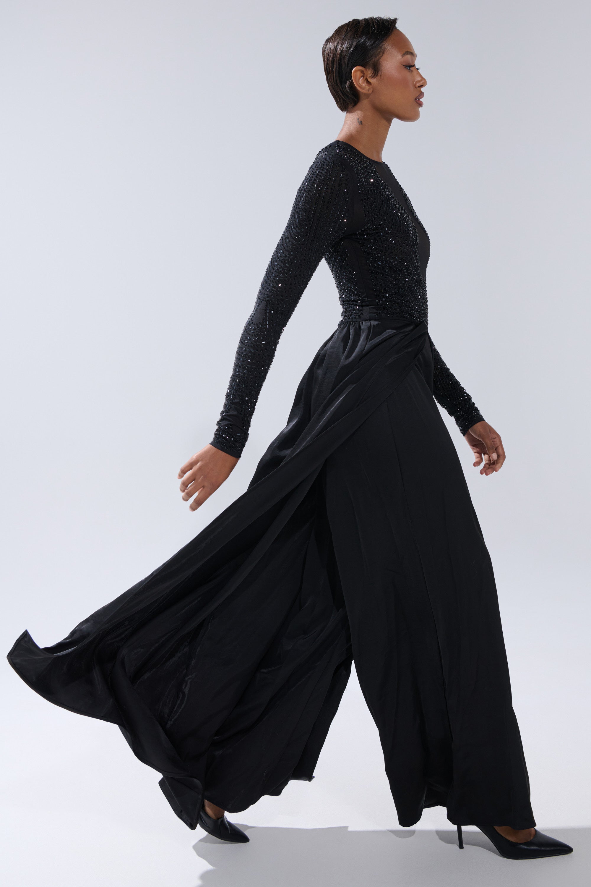 A woman in the MONACO SATIN MESH JUMPSUIT WITH RHINESTONES, featuring flowing wide-leg pants and subtle sparkle, walks in profile against a light gray background, finished with black high heels.