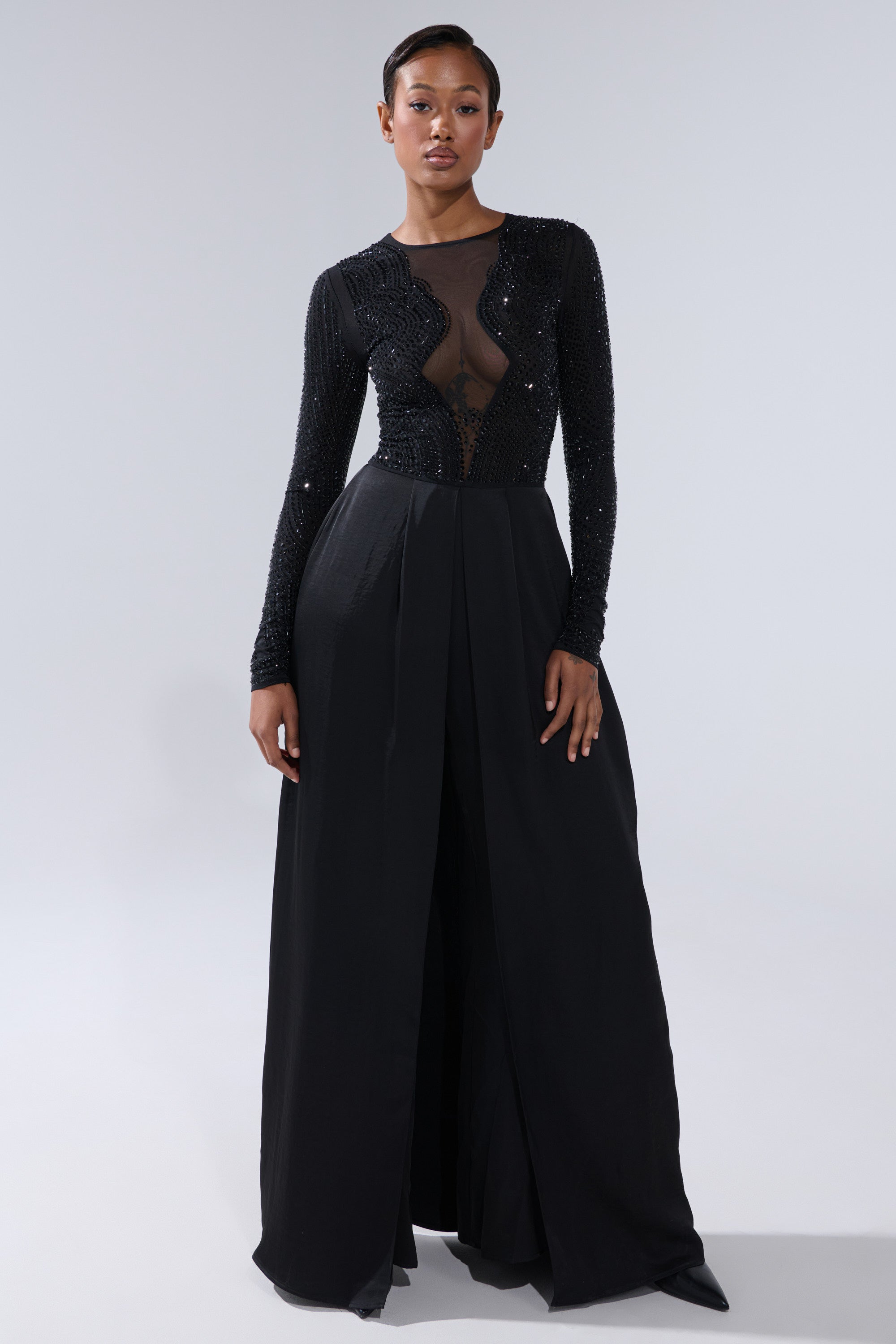 A woman poses in the MONACO SATIN MESH JUMPSUIT WITH RHINESTONES, featuring a sheer, rhinestone-embellished bodice and long sleeves. The sleek jumpsuit stands out against a plain, light gray background.