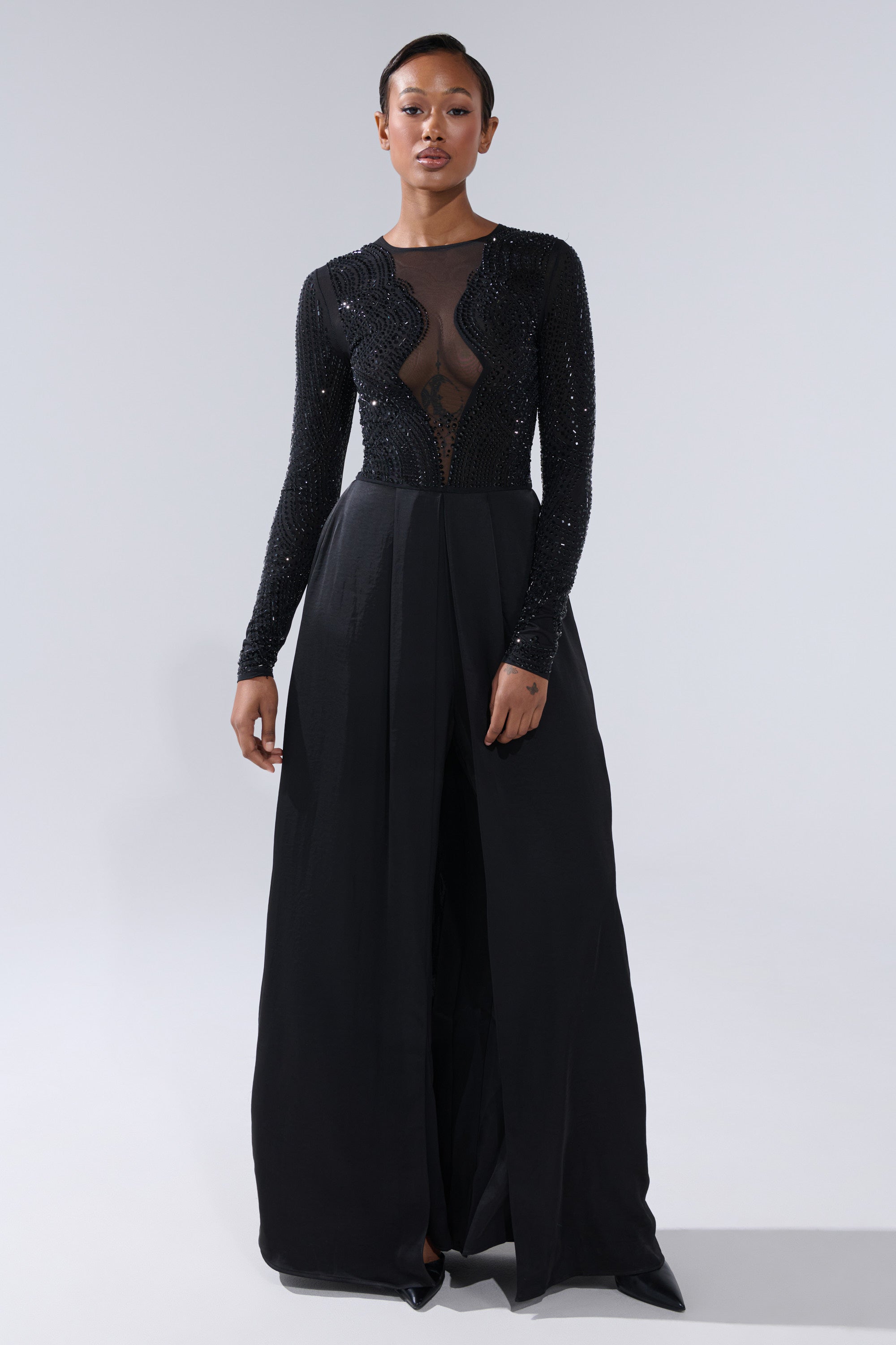 A woman faces forward in the MONACO SATIN MESH JUMPSUIT WITH RHINESTONES, featuring a sheer, rhinestone-embellished bodice and long sleeves. The jumpsuit creates an elegant maxi-length silhouette against a plain light gray background.