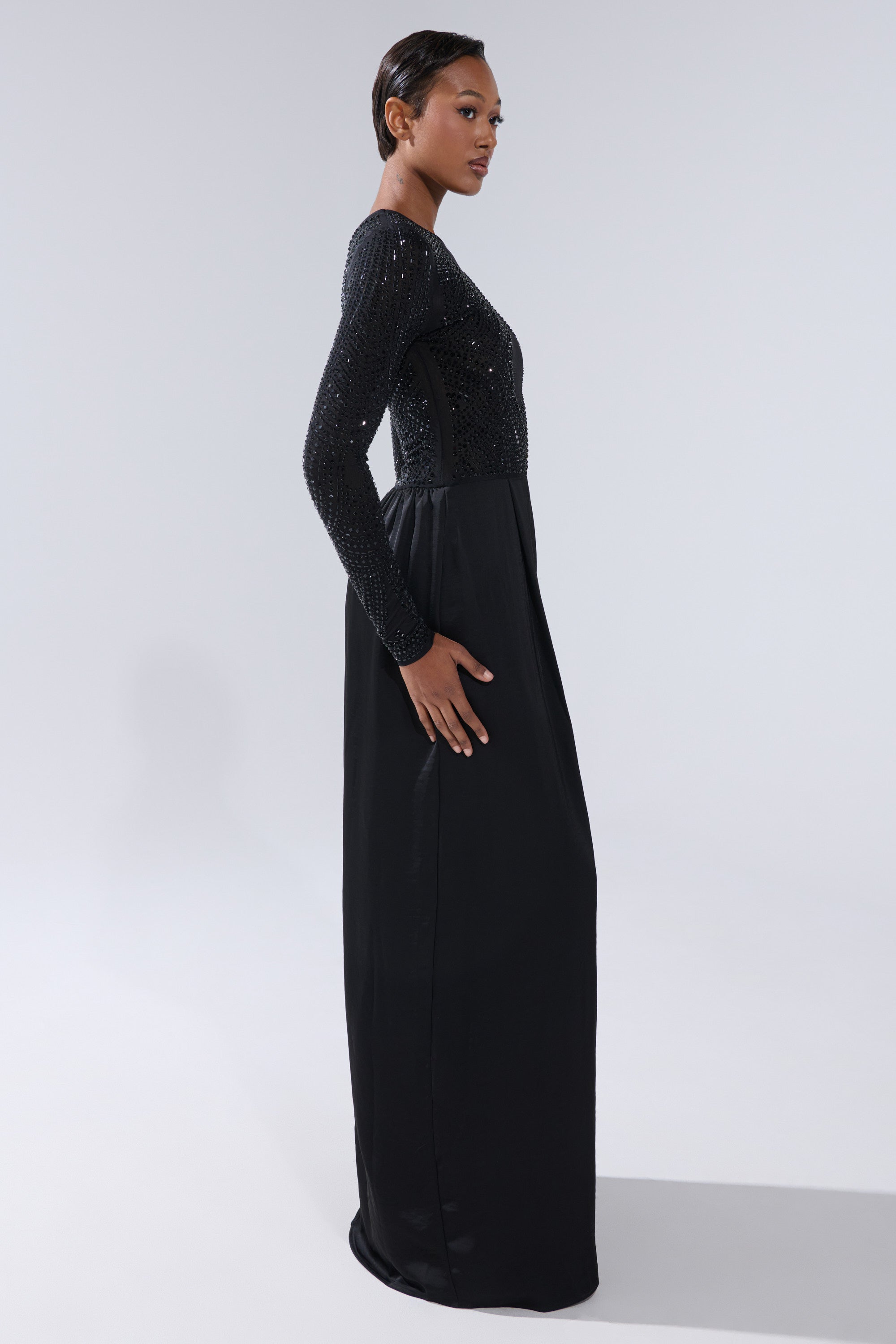 A woman stands in profile wearing the MONACO SATIN MESH JUMPSUIT WITH RHINESTONES, featuring a fitted, beaded bodice and long sleeves. The floor-length silhouette creates an elegant look as she poses against a light gray background.