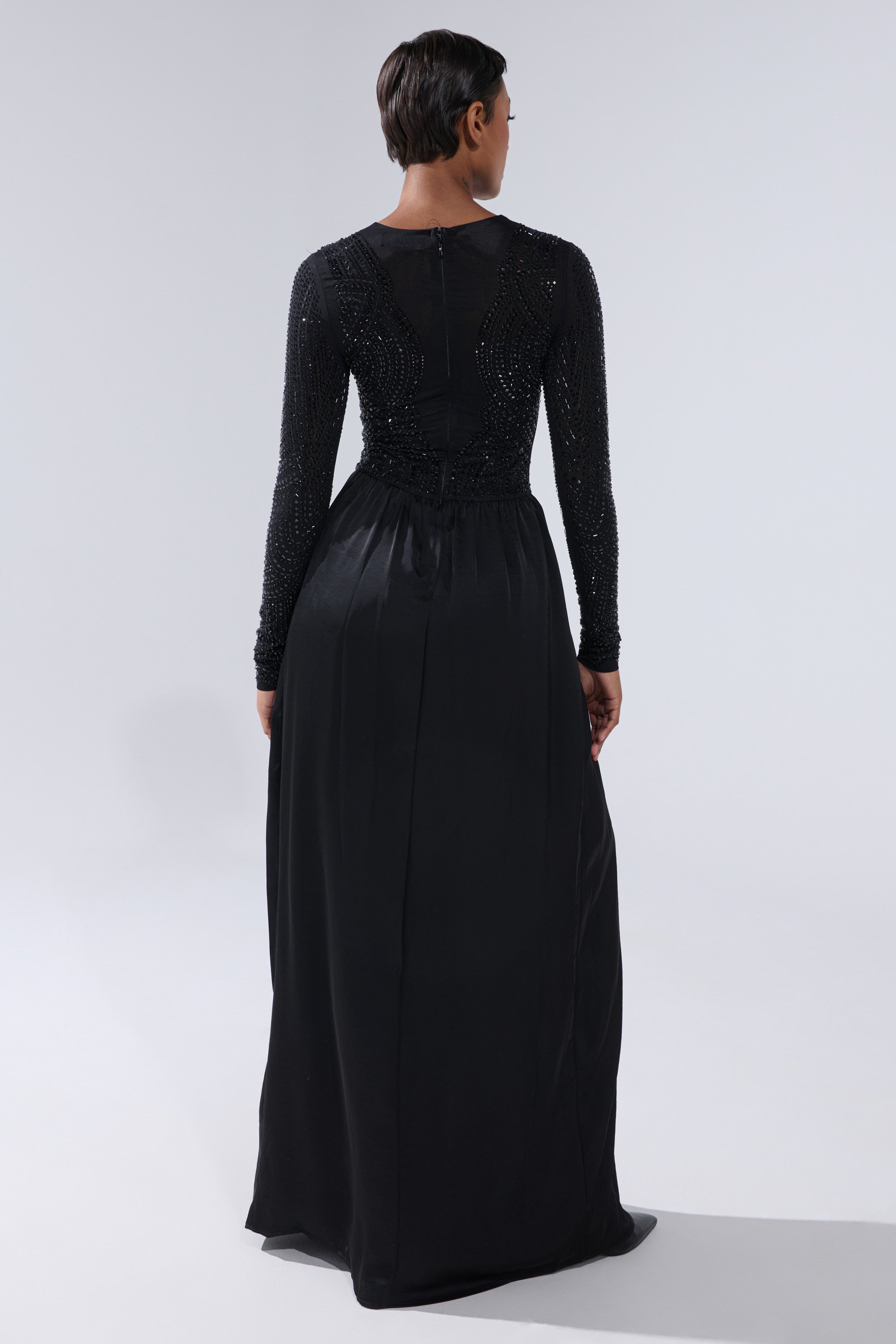 A person with short hair is shown from behind wearing the MONACO SATIN MESH JUMPSUIT WITH RHINESTONES, featuring bead detailing and a maxi silhouette, standing against a plain light gray background.