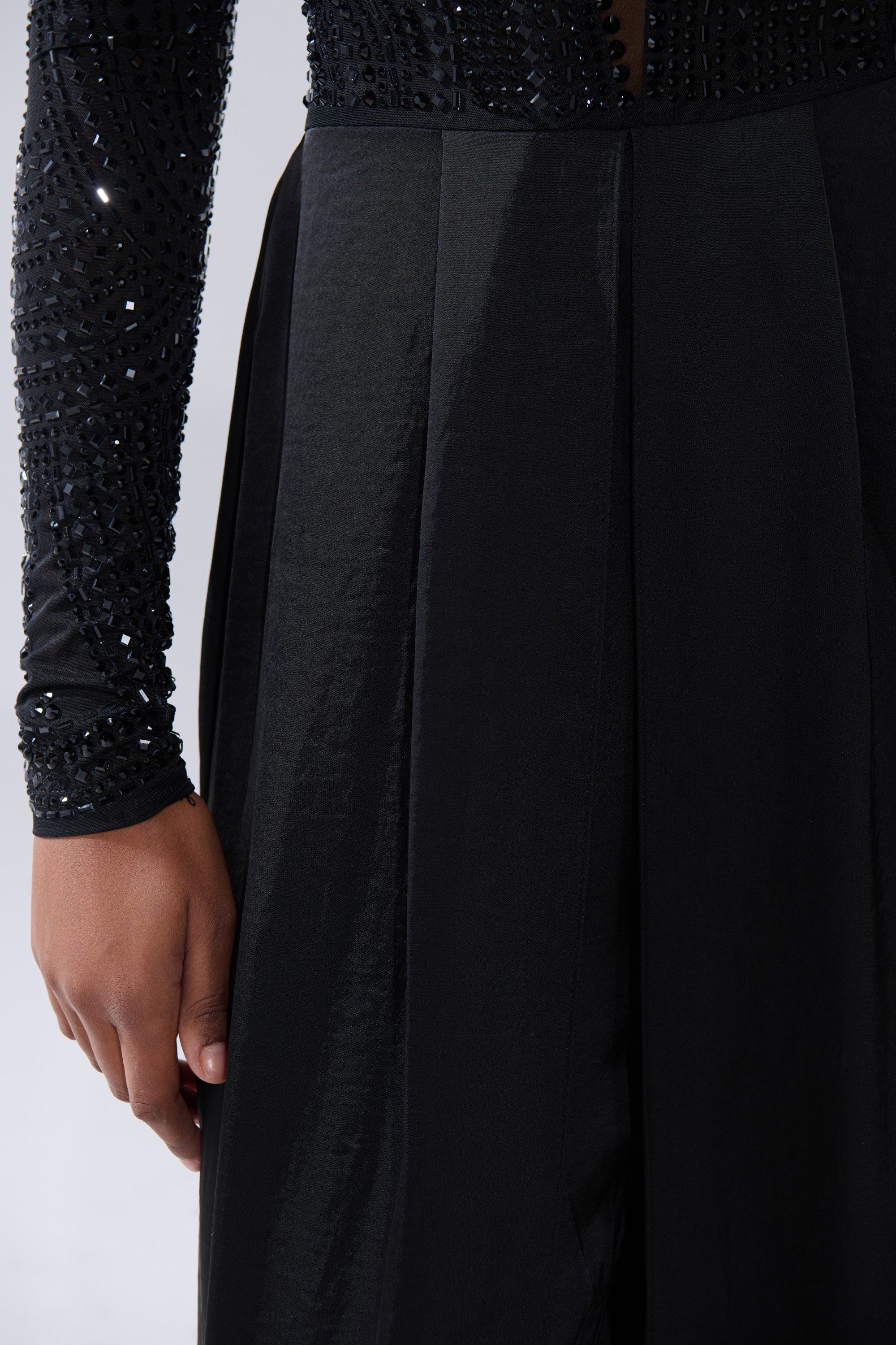 A close-up of a person wearing the MONACO SATIN MESH JUMPSUIT WITH RHINESTONES, featuring shiny embellishments, with their left arm at their side against a neutral background.