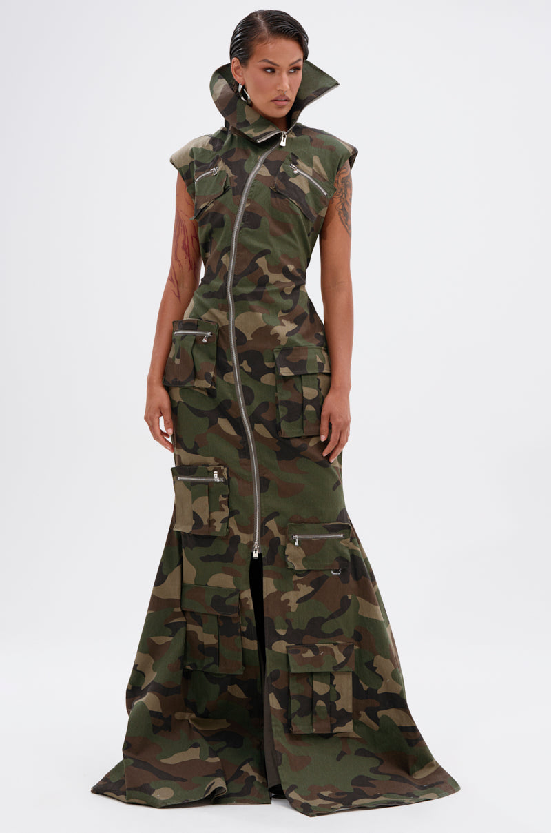 A person models the NOW YOU SEE ME CAMO PRINT MAXI DRESS, featuring a sleeveless design, high collar, cargo pockets, long front zipper, and flared hem. Slicked-back hair and visible arm tattoos complete the look.